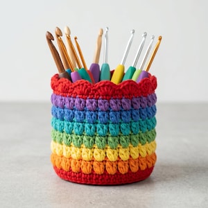 Op de afbeelding: Een regenbooggekleurde gehaakte container gevuld met verschillende haaknaalden. De container is cilindrisch met een geschulpte bovenrand. De haken zijn gemaakt van hout en metaal, met kleurrijke handvatten. De kleuren van de container lopen over van rood naar oranje, geel, groen, blauw en paars.