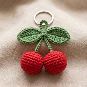 Puede incluir: Un llavero de ganchillo hecho a mano con dos cerezas rojas con hojas y tallos verdes. El llavero tiene un anillo de metal plateado. Las cerezas son redondas y texturizadas.