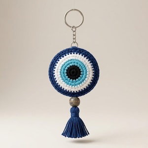 May include: A blue and white crocheted Evil Eye keychain with a silver key ring. The keychain has a dark blue tassel and a wooden bead. The Evil Eye design features concentric circles of black, light blue, white, and dark blue.