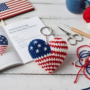 May include: A handmade crochet keychain in the shape of a heart, featuring the American flag design. The keychain is red, white, and blue, with white stars on a blue field. The image also shows a wooden American flag, crochet supplies, and a pattern.
