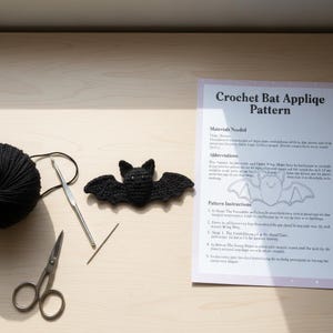 Pode incluir: Uma aplicação de morcego de crochê preto ao lado de uma bola de fio preto, uma agulha de crochê, tesouras, uma agulha e uma folha de padrão. A folha de padrão diz "Crochet Bat Appliqe Pattern".
