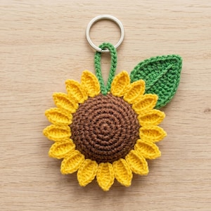 Puede incluir: Un llavero de girasol tejido a ganchillo con un centro marrón oscuro, pétalos de color amarillo brillante y una hoja verde única unida a una anilla de metal plateado. El girasol está hecho de hilo con un diseño circular y texturizado.