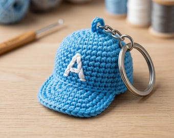 May include: A light blue crocheted baseball cap keychain with a white letter "A" on the front. The keychain has a silver ring and chain. The background includes spools of thread and a crochet hook.