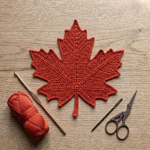 May include: A rust-coloured crocheted maple leaf applique positioned next to a matching ball of yarn, a metallic crochet hook, a long sewing needle, and a pair of ornate metal scissors. The items are arranged on a light-toned wooden surface.