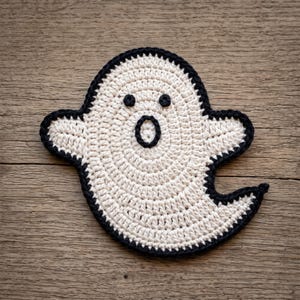 May include: A crochet ghost applique featuring an off-white body with a black yarn border. The ghost has two small black circular eyes and a larger black oval-shaped mouth centered on its face, all crafted with visible yarn stitches.