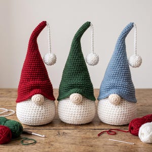 May include: Three handmade crochet gnomes with tall pointed hats in red, dark green, and light blue. Each gnome features a round beige nose, a white crocheted body, and a white pom-pom attached to the tip of the hat with a matching cord.
