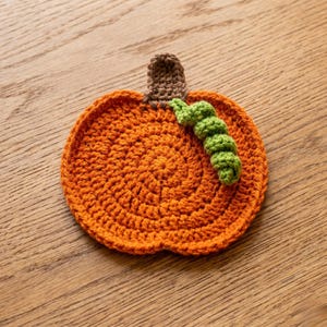 May include: Handmade orange crochet pumpkin coaster with a brown stem and green vine detail. This fall-themed coaster is perfect for adding a touch of autumn to your home decor. The coaster is approximately 5 inches in diameter.