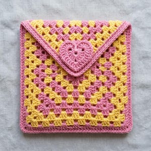 Pode incluir: Uma bolsa quadrada de crochê com padrão granny square em carreiras alternadas de fio rosa suave e amarelo brilhante. A aba estilo envelope é fixada com um motivo de coração tridimensional centralizado, feito em crochê com fio rosa.