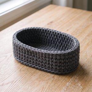 May include: An oval-shaped storage basket crafted from dark gray textile yarn. The basket features a consistent, dense crochet stitch pattern throughout its walls and base, creating a sturdy, open-top container with a uniform, textured appearance.