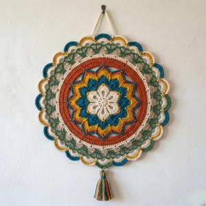 Puede incluir: Mandala redondo de ganchillo, colorido. El diseño presenta círculos concéntricos en tonos turquesa, naranja, amarillo, verde y crema. Una borla cuelga de la parte inferior. Se cuelga con un cordón color crema.
