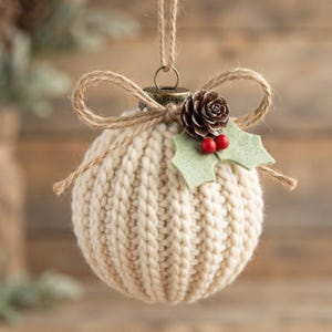 May include: A round Christmas ornament covered in a cream-colored ribbed knit fabric. The top features a jute twine bow, a small brown pinecone, two light green felt holly leaves, and two small red berries, all suspended by a jute twine loop.