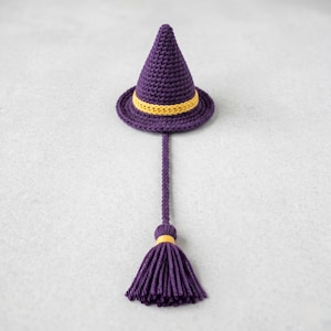 Puede incluir: Un sombrero de bruja morado tejido a ganchillo con una banda amarilla y una borla a juego. El sombrero tiene forma de cono con un ala ancha. La borla está unida al sombrero por un cordón largo y fino tejido a ganchillo. Es un artículo decorativo.