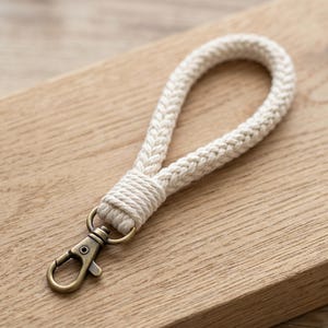 May include: Cream-colored woven wrist strap with a bronze-colored clasp. The strap is made of thick, braided cotton rope, looped to form a handle. The clasp is attached to a metal ring, and the strap is secured with wrapped cord.