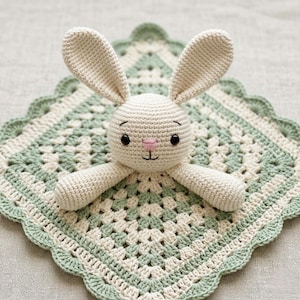 Peut inclure: Une couverture de sécurité au crochet avec une tête de lapin crème aux longues oreilles et au nez rose, fixée sur une couverture carrée à bords festonnés, réalisée en alternant du fil crème et vert sauge en mailles granny.