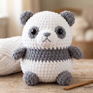 May include: A plush panda toy crafted from white and dark grey yarn. The stuffed animal features a round head with dark grey ears and eye patches, a white snout with a dark grey nose, and a round body with dark grey limbs and a white belly.