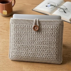 May include: A beige crocheted laptop sleeve with a button closure. The sleeve is made of a textured yarn and has a button made of wood. The laptop sleeve is on a wooden surface.