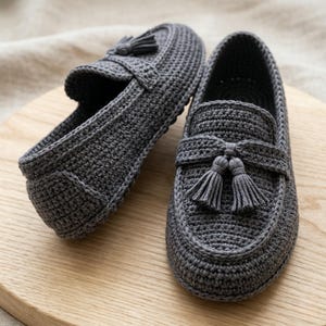 May include: A pair of grey crochet loafers with tassel detailing. The handmade shoes feature a textured stitch pattern and a classic loafer design. The loafers are displayed on a wooden surface.