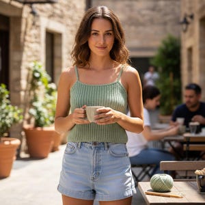 May include: A woman wearing a light green knitted tank top and light blue denim shorts, holding a beige mug. The tank top has thin straps and a ribbed texture. A ball of green yarn and a crochet hook sit on a wooden table.