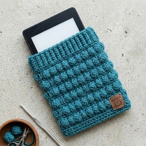 May include: Teal crochet e-reader cover with a textured bobble stitch design. The cover has a rectangular shape and a leather tag that reads "Saylabot" with a book icon. A crochet hook and small wooden bowl with yarn are also visible.