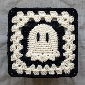 May include: A square, crocheted ghost design in black and off-white. The ghost has two vertical black eye slits. The square is framed with a black border and a floral-like pattern in off-white.