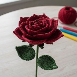 May include: A handmade, crocheted red rose with a green stem and leaves. The rose is in full bloom, with detailed petal work. A ball of red yarn and crochet hooks are in the background, suggesting a craft project. The rose is a decorative item.