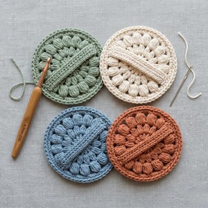 May include: Four handmade crochet coasters in shades of green, cream, blue, and rust. Each coaster features a circular design with a textured, raised pattern and a crocheted handle. A crochet hook and needle with thread are also present.