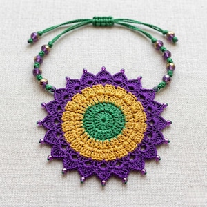 Può includere: Una collana con ciondolo circolare all'uncinetto caratterizzata da un cerchio centrale verde circondato da un anello giallo oro e uno strato esterno viola con bordi appuntiti. Il cordino verde include perline viola e oro e un nodo scorrevole.