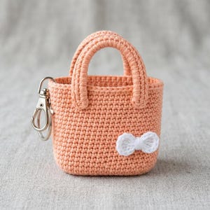 Op de afbeelding: Een perzikkleurige gehaakte mini-tas sleutelhanger met twee stevige handvatten en een kleine witte gehaakte strik aan de voorzijde. Een zilverkleurige metalen karabijnhaak is aan de zijkant van de tas bevestigd voor functioneel gebruik.