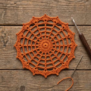 Pode incluir: Uma toalha de mesa de crochê laranja com um design de teia de aranha. A toalha de mesa circular é feita de fio e tem um padrão de trabalho aberto detalhado. Uma agulha de crochê e uma agulha são visíveis em uma superfície de madeira.