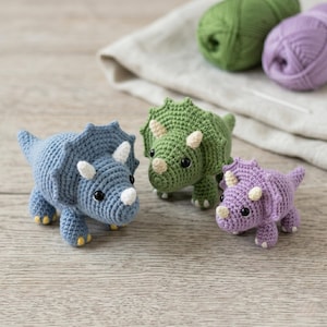 May include: Three crocheted triceratops dinosaurs in blue, green, and purple. Each has white horns and accents, with black eyes and yellow feet. The dinosaurs are handmade and sit on a wooden surface.