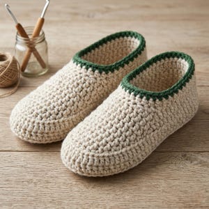 Puede incluir: Un par de zapatillas de ganchillo hechas a mano, de color crema, con un ribete verde oscuro. Las zapatillas están hechas de un hilo grueso y tienen un aspecto texturizado. También se ven ganchillos e hilo.