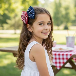Pode incluir: Uma menina usa um laço de cabelo vermelho, branco e azul, que lembra a bandeira americana. O laço é feito de material de malha. A menina está vestindo um vestido branco e sorrindo. O fundo está desfocado.