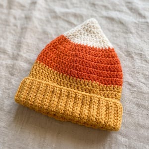May include: A crochet beanie hat featuring a color-blocked design with a white pointed top, a wide orange middle section, and a yellow ribbed brim. The hat is constructed with visible yarn stitches in a triangular shape resembling a candy corn.