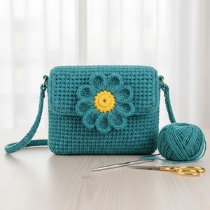 May include: Teal crochet crossbody bag with a large teal and yellow flower applique. The bag has a flap closure and a long strap. A ball of teal yarn, a crochet hook, and scissors are next to the bag.