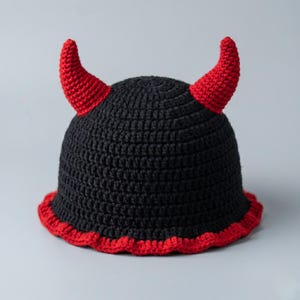 May include: A black crochet hat with red trim and two red horns. The hat has a rounded crown and a scalloped red edge. The horns are curved and point upwards. The hat is likely handmade and suitable for costume or novelty wear.