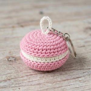 May include: A crocheted macaron keyring featuring two pink textured biscuit halves with a cream-coloured filling in the centre. A small white crocheted loop is attached to the top, connected to a silver-toned metal split ring for keys.