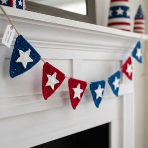 May include: A handmade crochet banner with alternating red and blue triangle flags, each featuring a white star. The banner is strung on twine and displayed on a white mantelpiece, with additional star-themed decorations in the background.