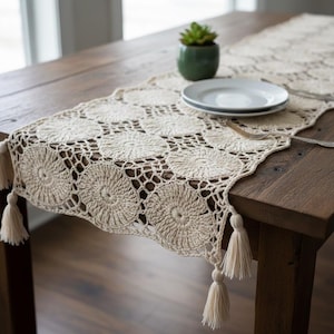 May include: A cream-coloured crochet table runner with a floral pattern and tassels. The runner is placed on a wooden table. A small green potted plant and two white plates are visible in the background. The runner's design features repeating circular motifs.