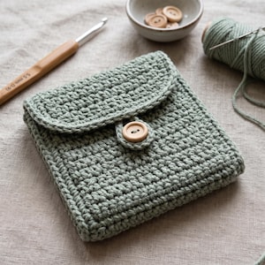 Può includere: Una pochette quadrata all'uncinetto color verde salvia con chiusura a patta fissata da un bottone in legno marrone chiaro. La pochette presenta un motivo a maglia fitta ed è accompagnata da un uncinetto da 5,5 mm, una ciotola di bottoni in legno e un gomitolo di filato abbinato.