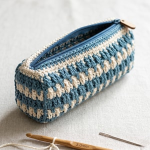 May include: A handmade crochet pencil case in blue and cream. The rectangular case features a zipper closure and a wooden zipper pull. A crochet hook and needle are visible near the case.