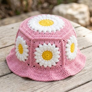 May include: A pink crochet bucket hat with white and yellow daisy motifs. The hat has a wide brim and is made of a soft, textured yarn. The daisy design is repeated around the hat, a charming summer accessory.