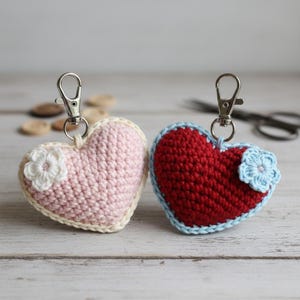 May include: Two handmade crochet heart keychains. One is pink with a white flower and cream trim, the other is red with a light blue trim and flower. Both have silver-colored metal clasps.