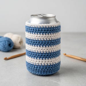 Puede incluir: Un portabotellas de ganchillo a rayas azules y blancas. El portabotellas está envuelto alrededor de una lata de aluminio plateada. El patrón de ganchillo presenta rayas horizontales azules y blancas. Se ven ganchos de ganchillo y lana en el fondo.
