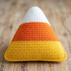 May include: A handmade, crocheted candy corn pillow. The pillow is triangular, with white, orange, and yellow horizontal stripes. The pillow is sitting on a wooden surface.