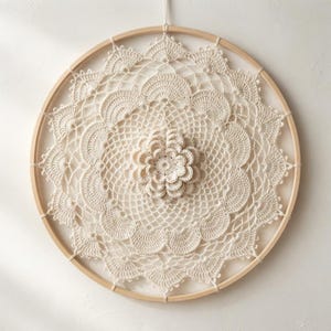 May include: A circular wall hanging featuring an off-white crocheted doily stretched within a light-colored wooden hoop. The intricate lace pattern radiates from a central three-dimensional crocheted flower, creating a detailed, layered design.