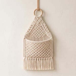 Puede incluir: Un tapiz de macramé color crema que presenta un bolsillo central con un patrón de nudos en forma de diamante. La pieza está suspendida de un anillo de madera lisa de tono claro y terminada con una fila de flecos verticales y rectos.