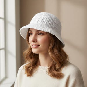 May include: A white crochet bucket hat with a small brim. The hat is made of a textured, open-weave material. The hat is worn by a person with long, wavy, light brown hair and a cream-colored sweater.