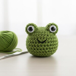 May include: A handmade green crochet frog with large googly eyes and a stitched smile. A ball of green yarn and a crochet hook are in the background. This whimsical item is perfect for a child's toy or a unique home decor piece.