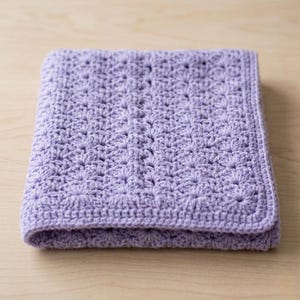 Może przedstawiać: Złożony, lawendowy koc szydełkowy. Koc ma wzorzystą fakturę i wykończoną krawędź. Miękki, ręcznie robiony koc jest wykonany z przędzy i jest starannie złożony na jasnej drewnianej powierzchni.