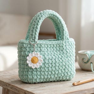 May include: A mint green, hand-crocheted tote bag with a matching handle. A daisy-shaped charm with a yellow center and white petals hangs from the handle. The bag sits on a wooden surface.